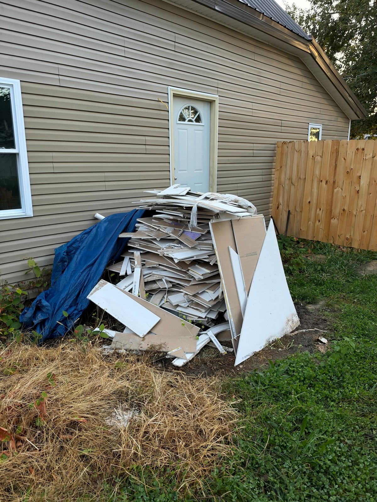 Pile of discarded construction debris and white boards covered with blue tarp outside a residential house
