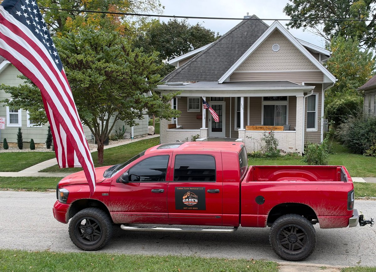 Red pickup truck with American flag parked in front of gray house with landscaping