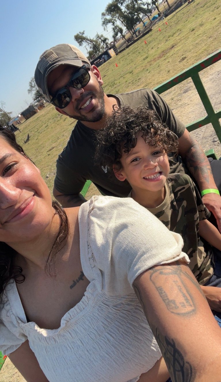 Three people smiling for a selfie at an outdoor park with green grass and trees in the background on a sunny day.